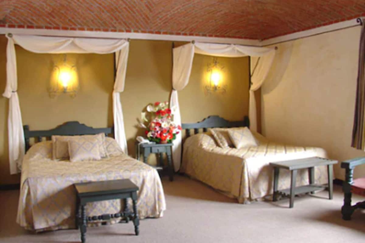 A room with two beds, a bench, a nightstand, and a wall-mounted lamp.