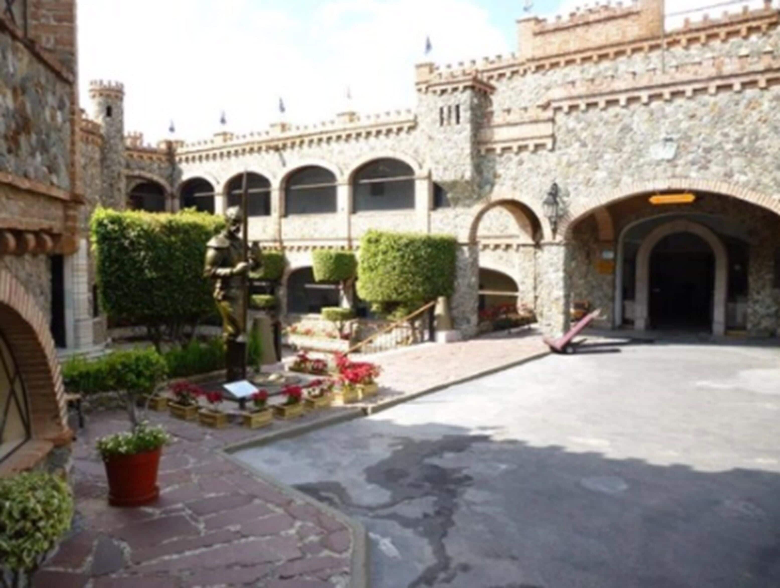 A courtyard with a statue, stone buildings with arches, and potted plants.