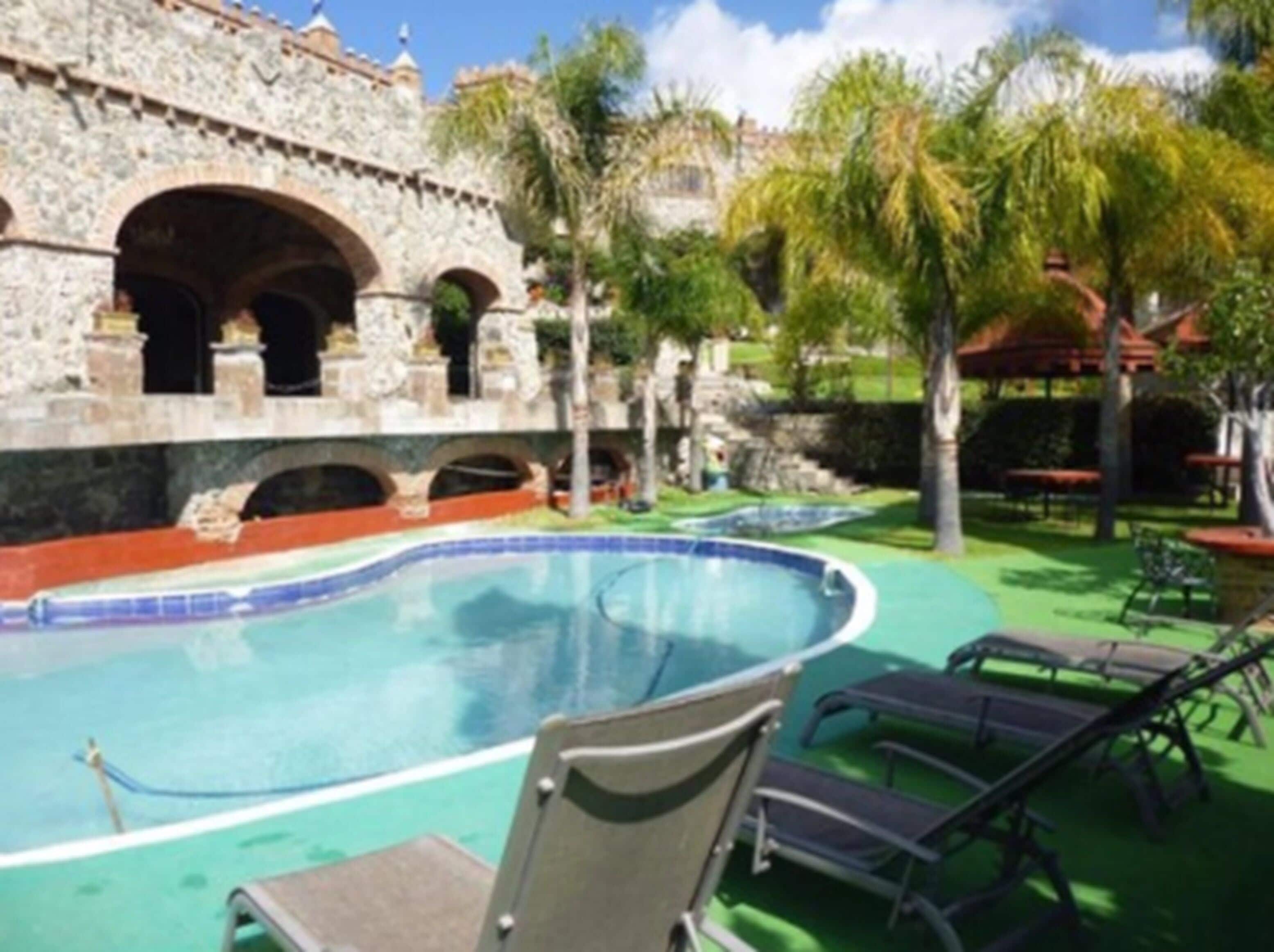 A swimming pool with lounge chairs, a stone building with arches, and palm trees.