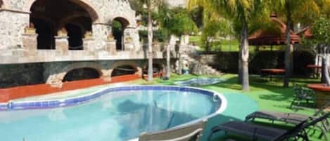 A swimming pool with lounge chairs, a stone building with arches, and palm trees.