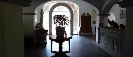 Un couloir faiblement éclairé, avec une porte en arc donnant sur une pièce plus lumineuse. On aperçoit un pot de plante près de la porte et une table avec une chaise au premier plan.