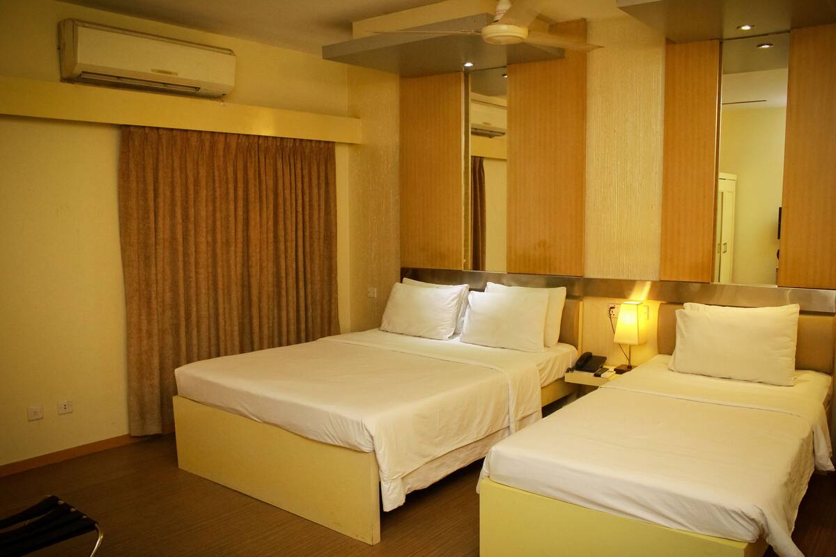 A hotel room with two beds, a ceiling fan, an air conditioning unit, a bedside table with a lamp, and a mirror.