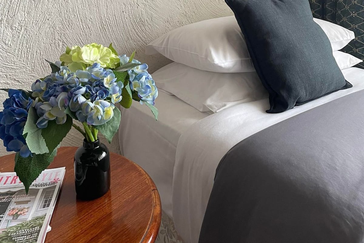 A hotel room with a bed, pillows, a vase with flowers, and a magazine on a wooden table.