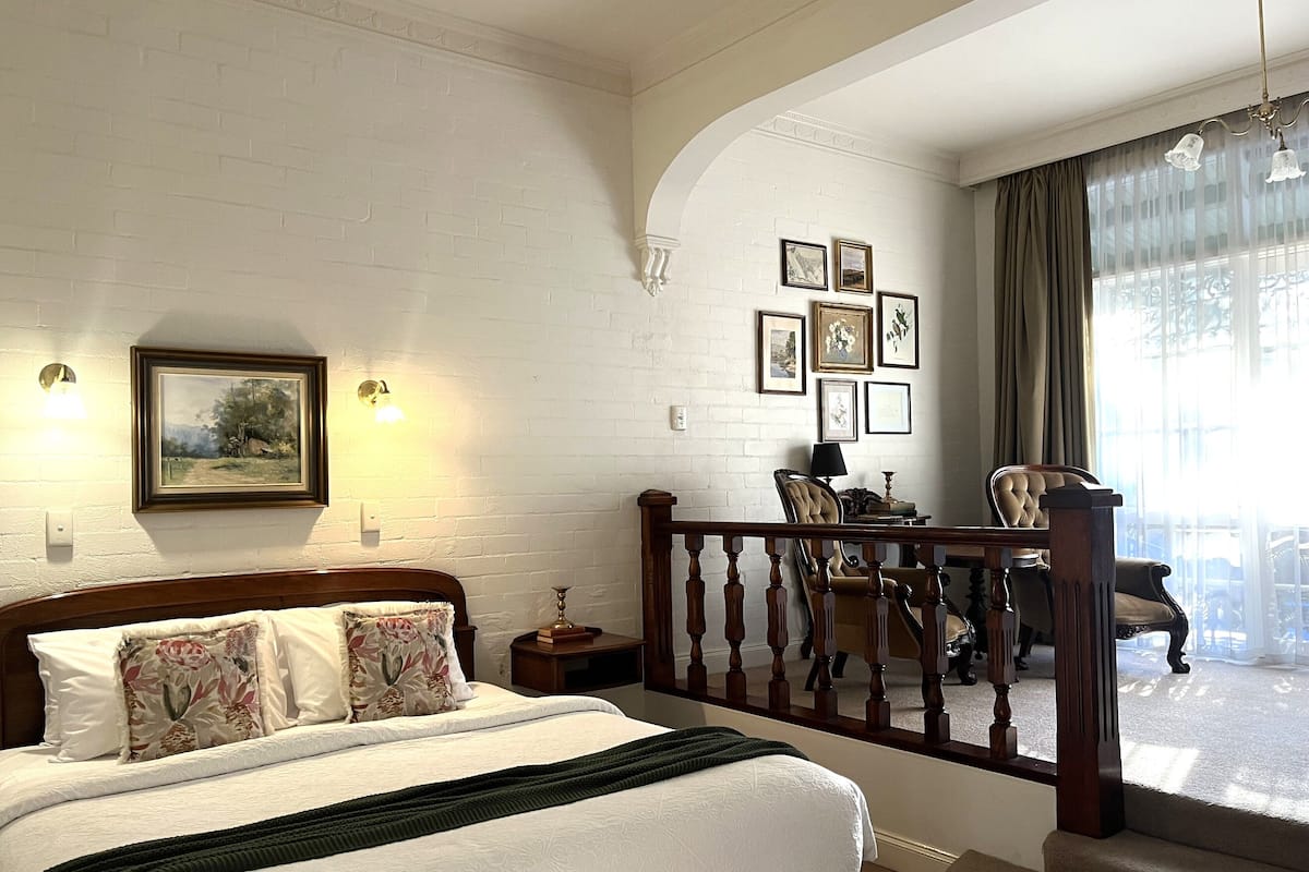 A bedroom with a large bed, a wooden headboard, a nightstand, a lamp, and a chair. There are framed pictures on the wall and a balcony with a railing.