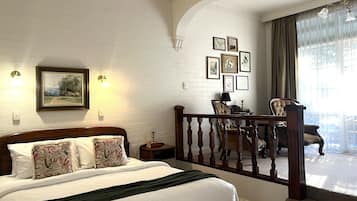 A bedroom with a large bed, a wooden headboard, a nightstand, a lamp, and a chair. There are framed pictures on the wall and a balcony with a railing.