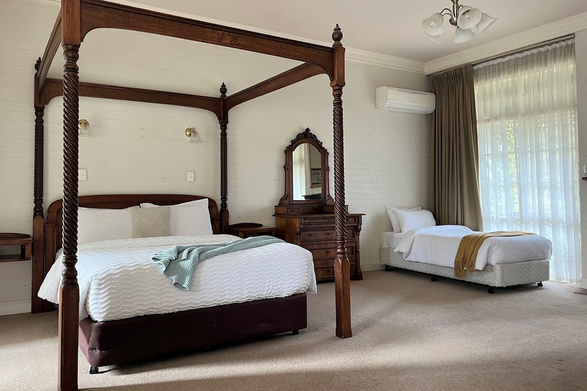 A four-poster bed with a wooden frame, a bedside table, a dresser, a mirror, and a window with curtains.