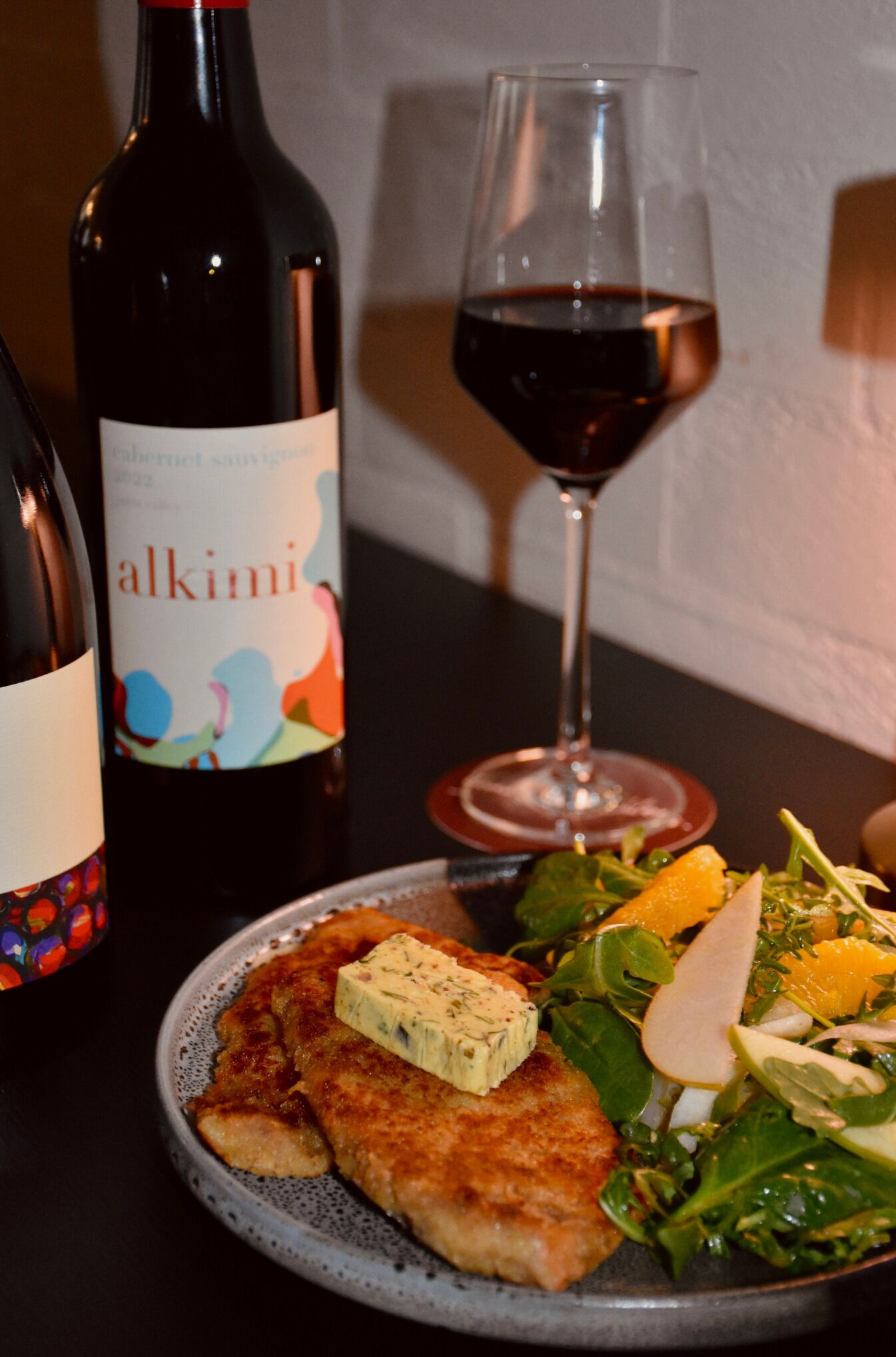 A plate with a piece of meat, a glass of red wine, and a bottle of wine with a colorful label.