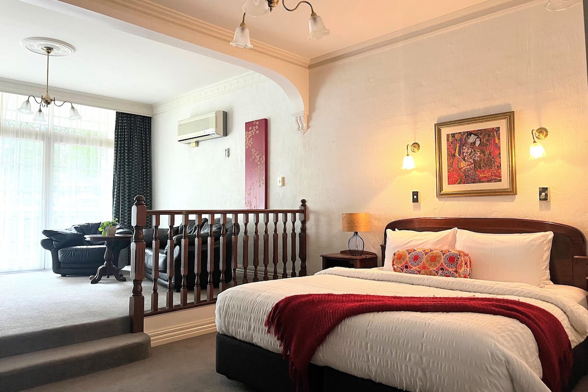 A bedroom with a large bed, a wooden headboard, a red throw blanket, and a nightstand with a lamp. There is a staircase leading to an outdoor area with a view of a building.