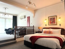 Quarto com uma cama grande, cabeceira de madeira, manta vermelha e mesa de cabeceira com luminária. Escada leva a área externa com vista para um edifício.