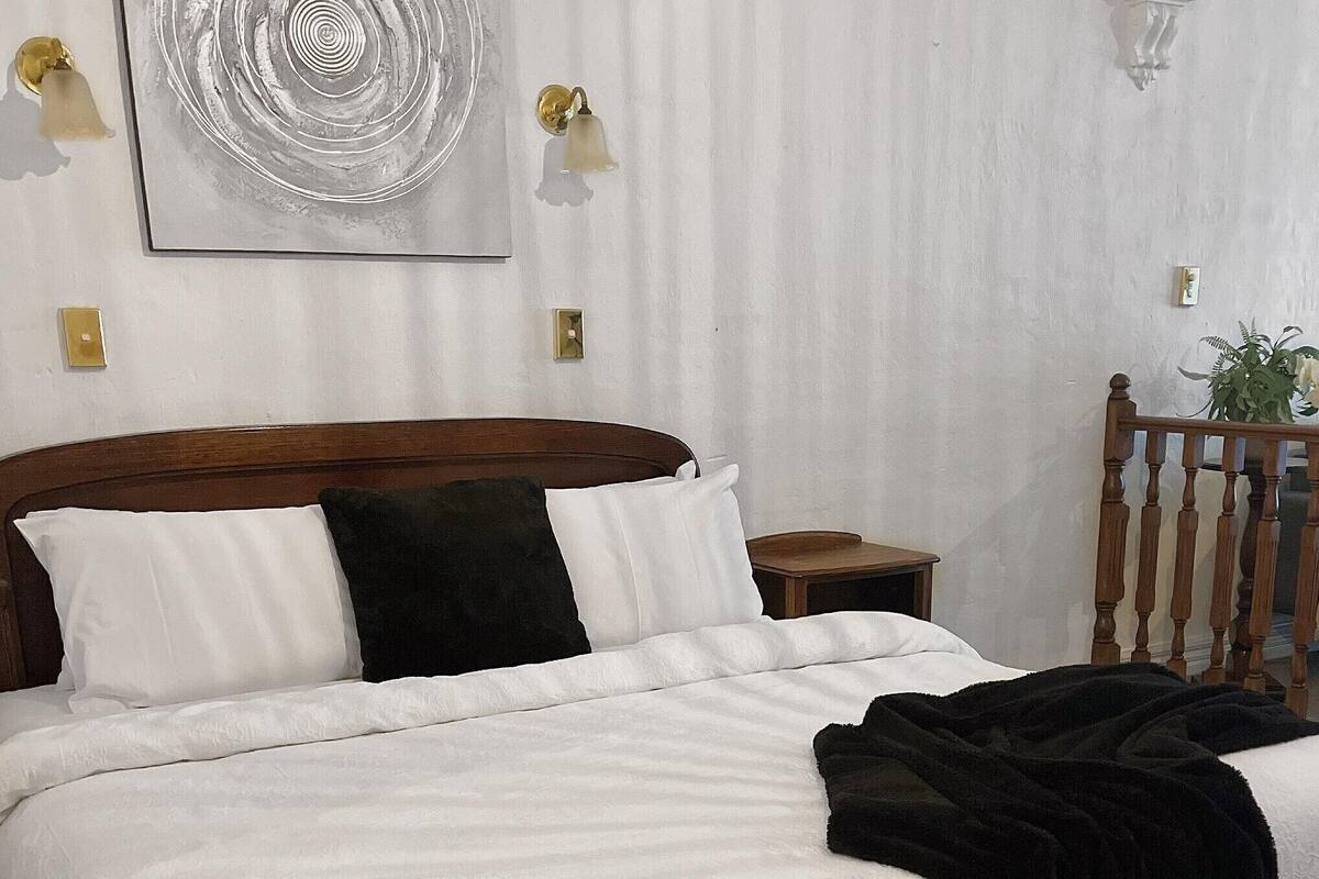 A neatly made bed with white and black pillows, a wooden headboard, a bedside table, and a wall-mounted light fixture.