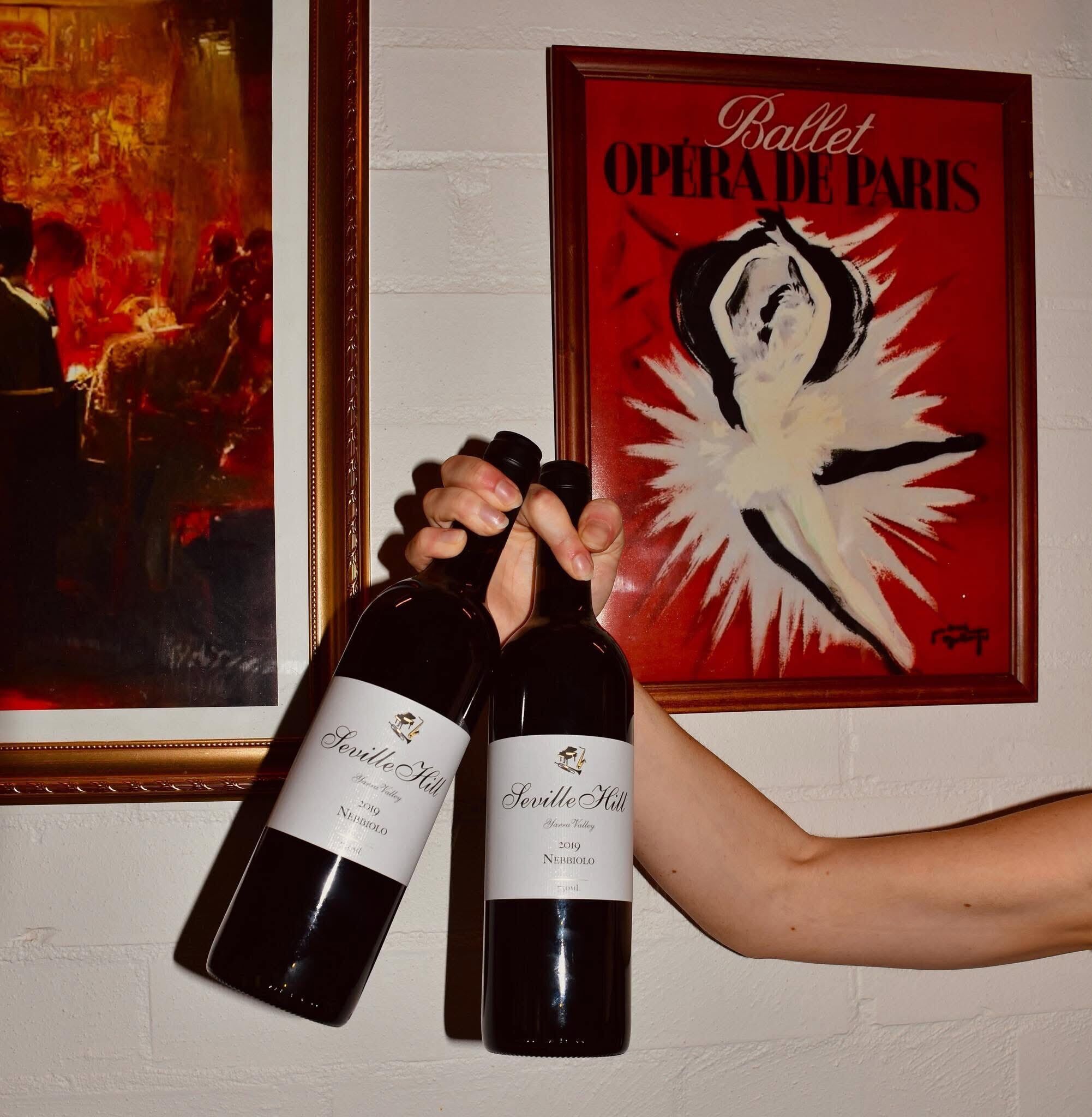 Two bottles of wine with labels, a framed painting, and a poster on a wall.