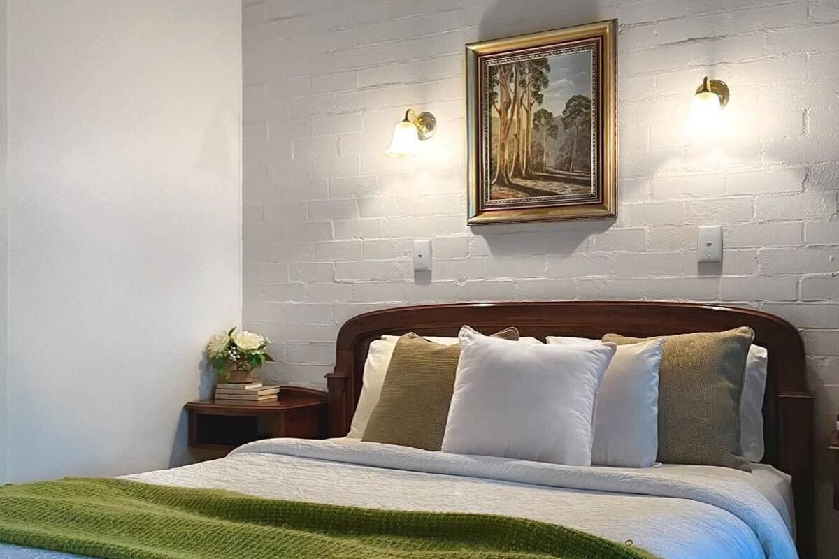 A bed with a green blanket, a nightstand with a vase of flowers, and a framed picture on the wall.