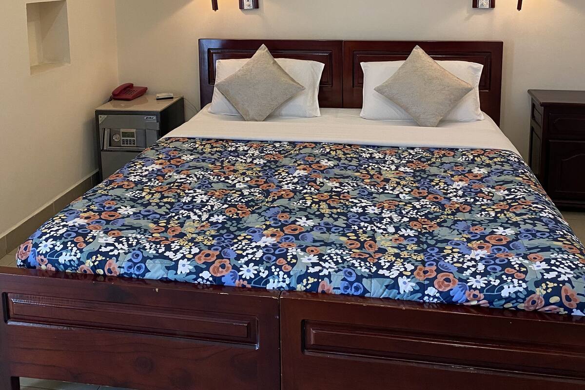 Un lit bien fait, recouvert d’une couvre-lit à motifs floraux, deux lampes de chevet, une œuvre d’art encadrée au-dessus du lit et une petite table de chevet avec un téléphone.
