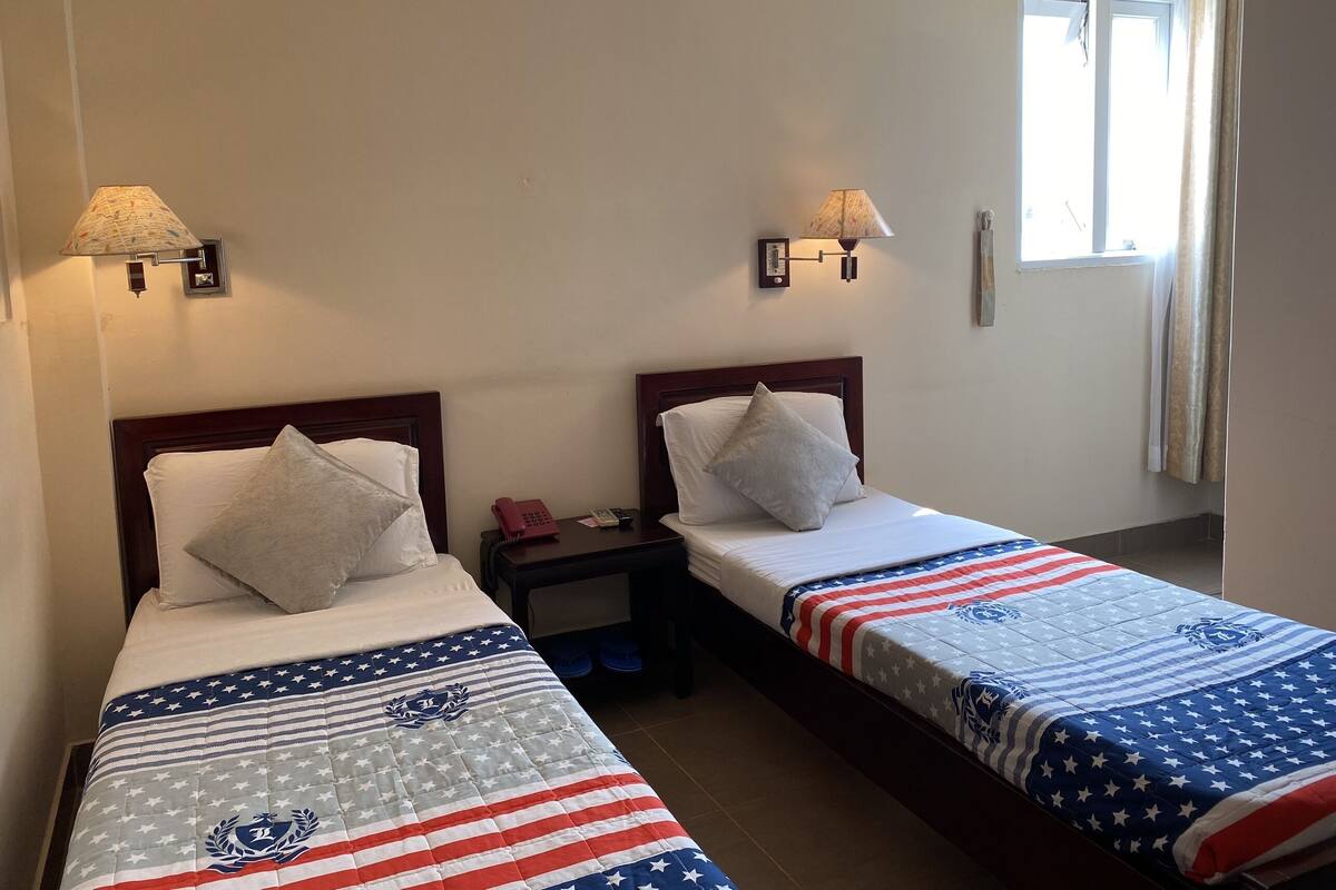 Une chambre d’hôtel avec deux lits, chacun recouvert d’une courtepointe matelassée ornée d’un motif patriotique. Des lampes murales sont fixées de chaque côté des lits, et une fenêtre avec des rideaux se trouve sur le côté droit.