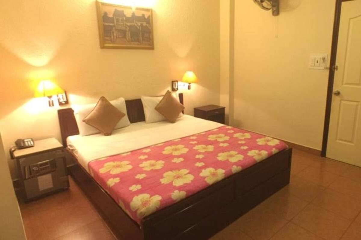 Une chambre d’hôtel comprenant un lit, deux tables de chevet, une commode, une lampe fixée au mur, une porte et un tableau encadré accroché au mur.