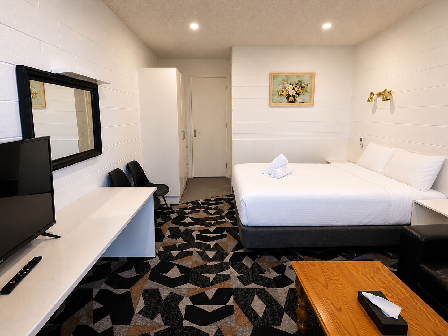 A hotel room with a bed, a desk with a TV, a chair, a wooden table, and a patterned rug.