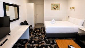 A hotel room with a bed, a desk with a TV, a chair, a wooden table, and a patterned rug. - Pathfinder Motel (Kew)