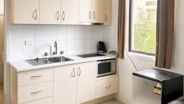 A compact kitchen with a sink, stove, microwave, and a small refrigerator. There is a window with curtains and a view of the outside. - Pathfinder Motel (Kew)