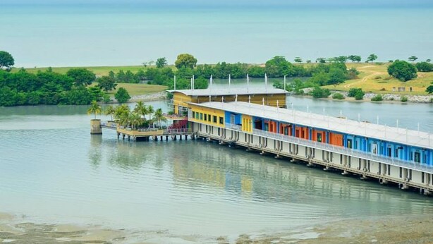 Un muelle largo y colorido se extiende hacia una masa de agua tranquila, con un edificio al final que cuenta con varias habitaciones.