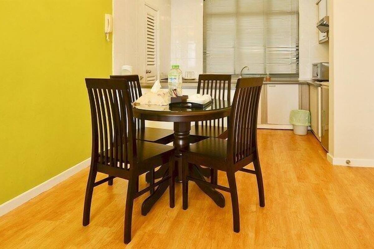 A dining area with a round table and four chairs, a window with blinds, a kitchen area with appliances, and a trash can.