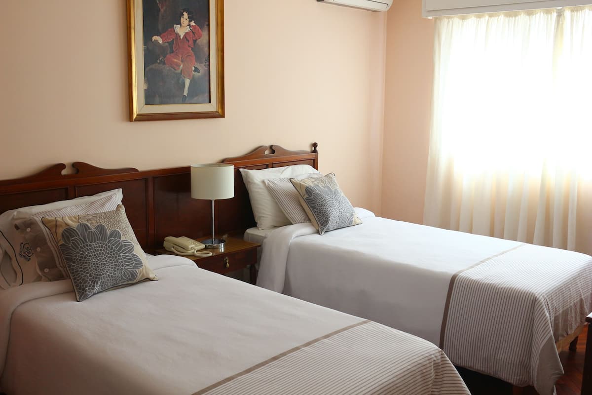 A hotel room with two beds, a bedside table with a lamp, a painting on the wall, and a window with curtains.