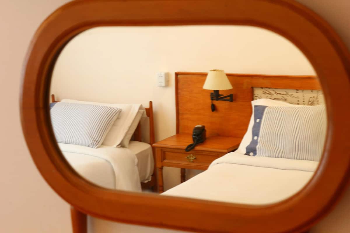 A mirror reflecting a hotel room with two beds, a nightstand with a lamp, and a telephone.