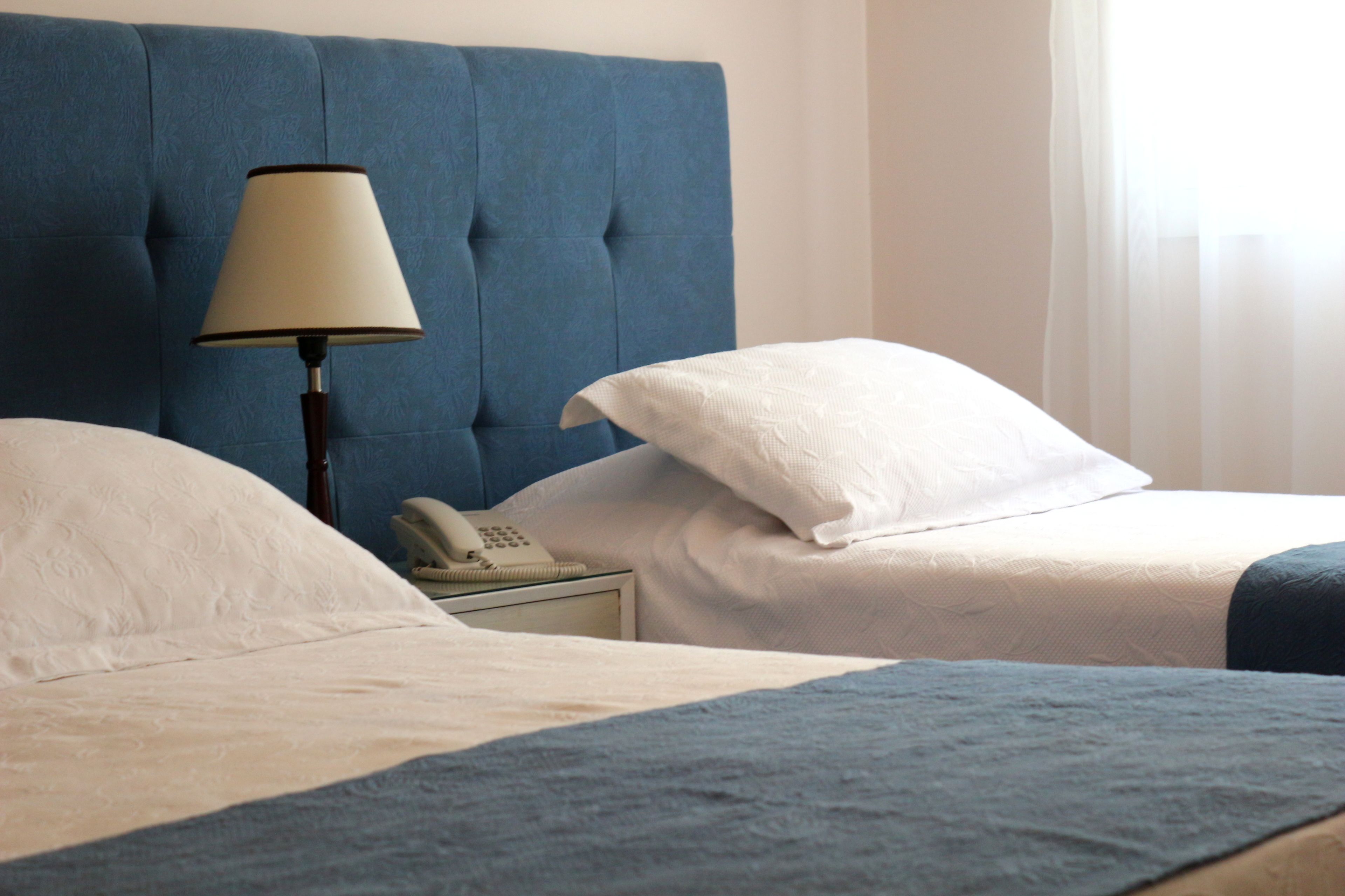 Une chambre d’hôtel avec une tête de lit capitonnée, une lampe de chevet, un téléphone et des lits parfaitement faits, recouverts de draps blancs et bleus.