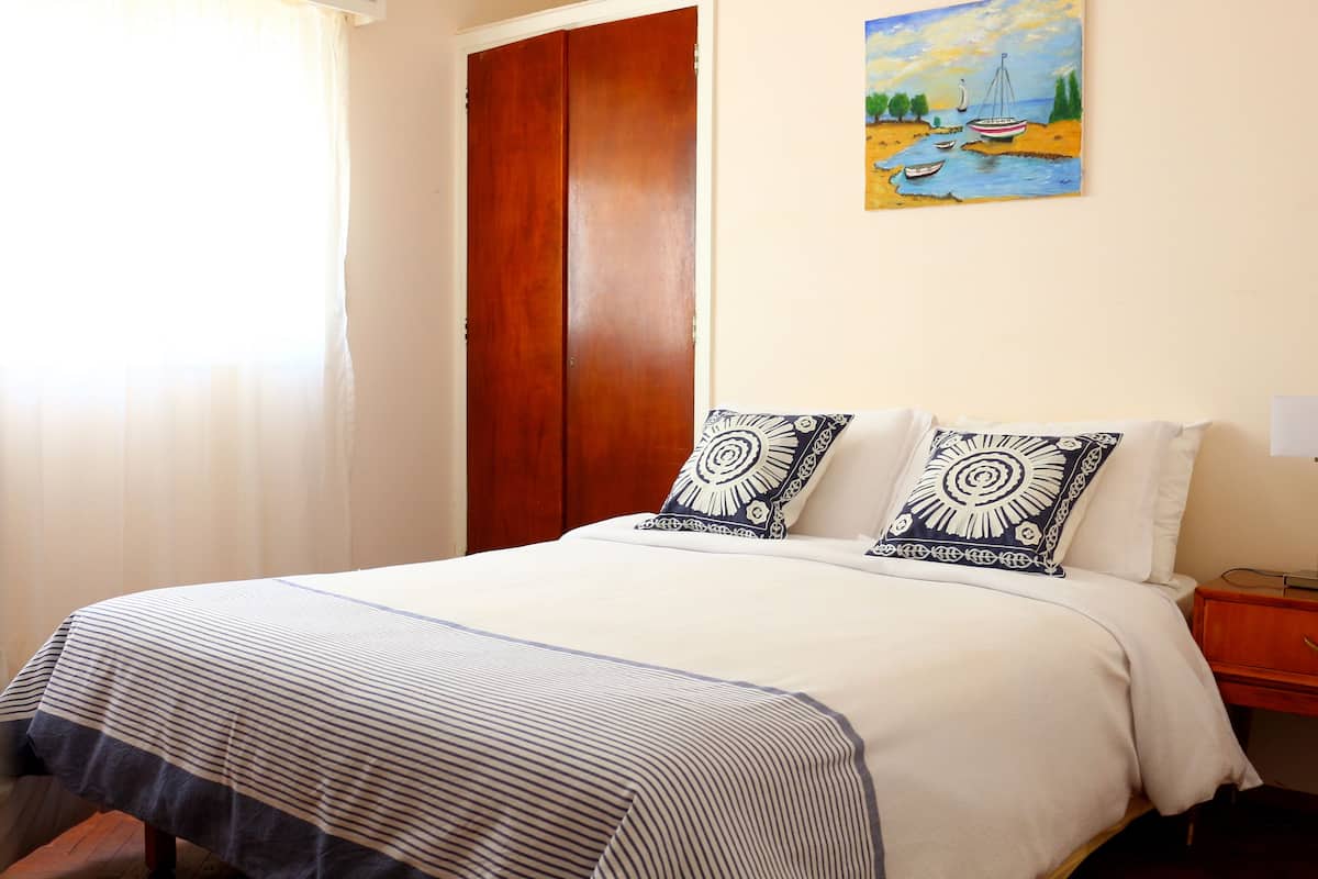 A neatly made bed with a striped bedspread and two patterned pillows, a wooden wardrobe, and a painting of a river scene on the wall.