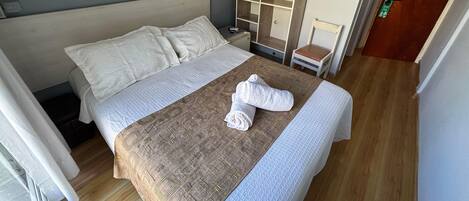 A neatly made bed with a patterned bedspread and pillows, a wooden floor, a small bedside table with a lamp, and a mirror on the wall.