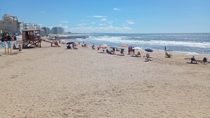 A beach with people sunbathing and walking along the shore, a lifeguard tower, and buildings in the background. - Hotel Playa Brava (Punta del Este)