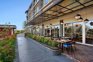 An outdoor dining area with tables and chairs, a covered walkway, and a building with large windows in the background. - Azana Boutique Hotel Denpasar (Denpasar)