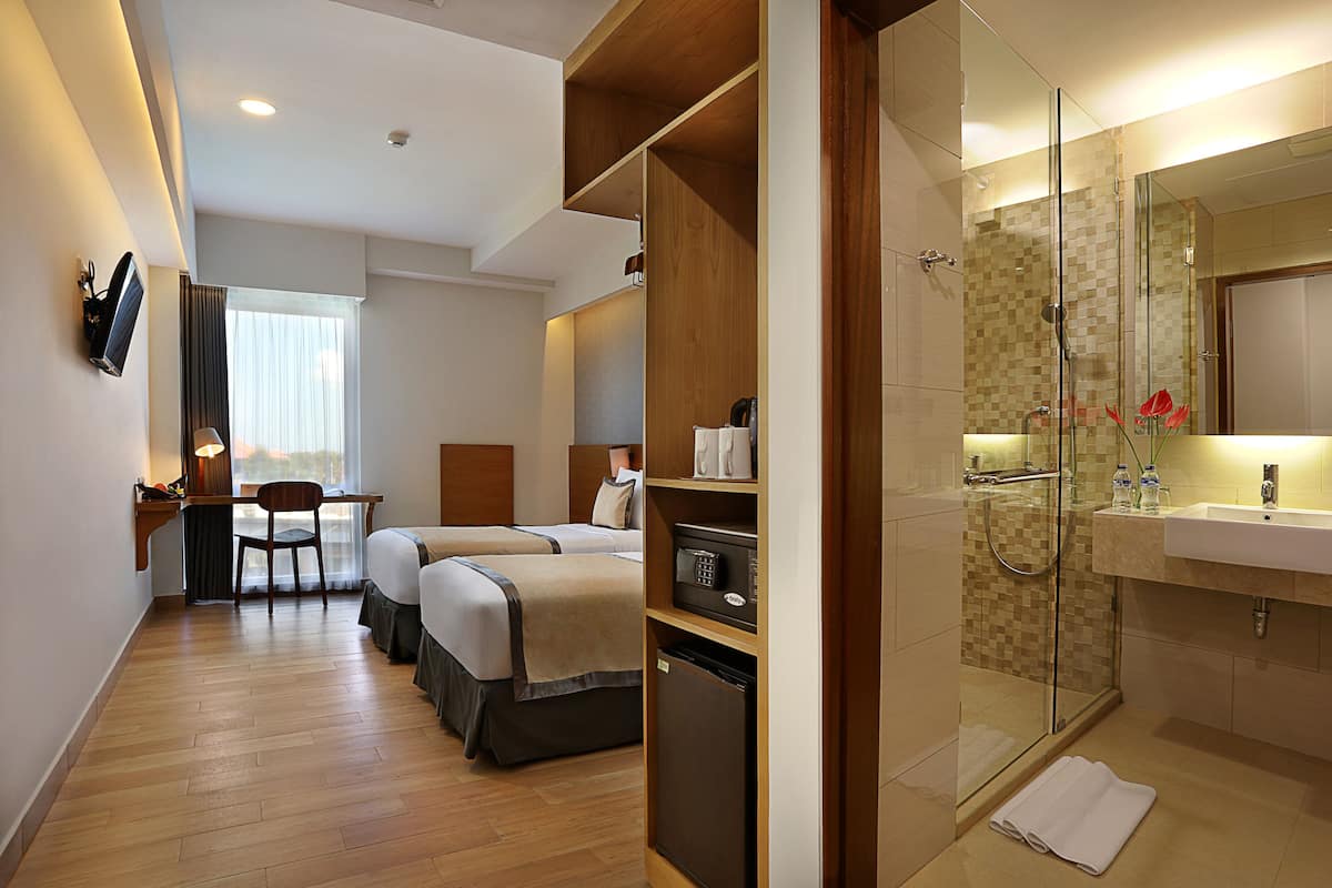 A hotel room with a desk, two beds, a mini-fridge, a shower area, and a TV mounted on the wall.