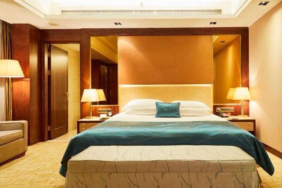 A hotel room with a large bed, two bedside tables with lamps, a nightstand, a sofa, and a door leading to another room.