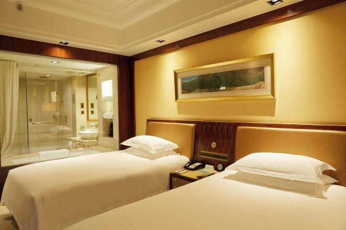 A hotel room with two beds, a large mirror, a painting on the wall, and a bathroom visible through the glass door.
