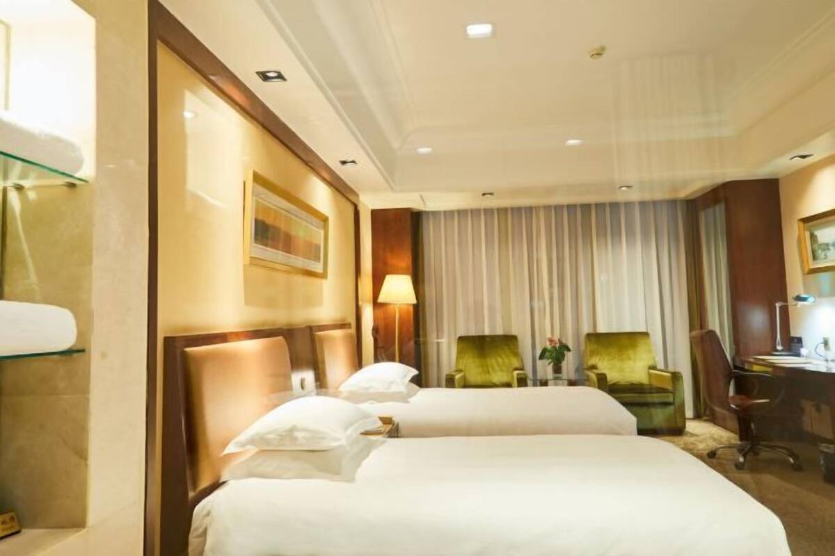 A hotel room with two beds, a desk, two chairs, a lamp, and a window with curtains.