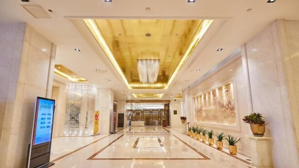 A spacious hotel lobby with a modern design, featuring a central chandelier, a digital information kiosk, and a row of potted plants along the walls.