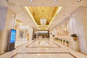 A spacious hotel lobby with a modern design, featuring a central chandelier, a digital information kiosk, and a row of potted plants along the walls.