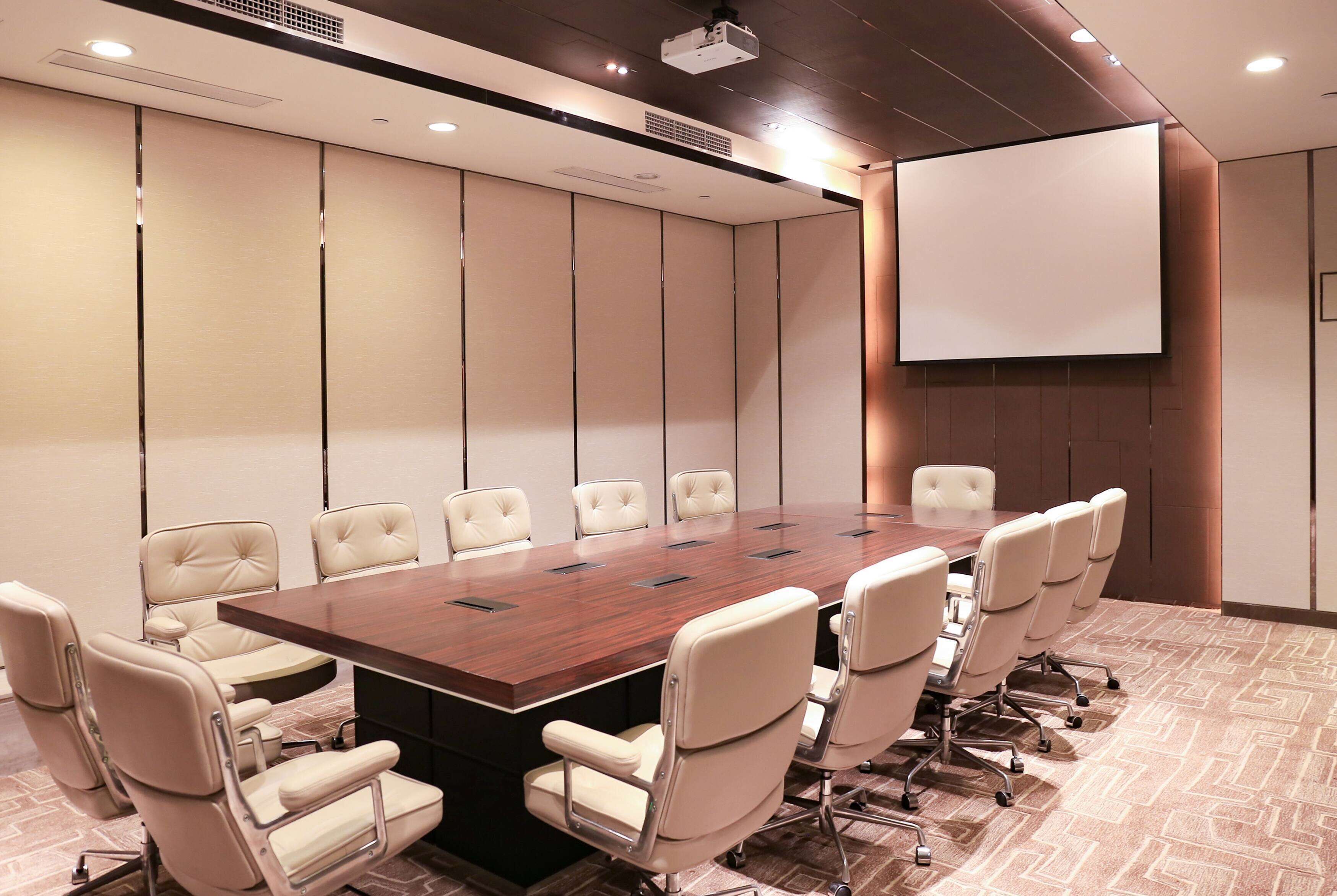 A conference room with a large wooden table, beige upholstered chairs, a projector screen, and a tiled floor.