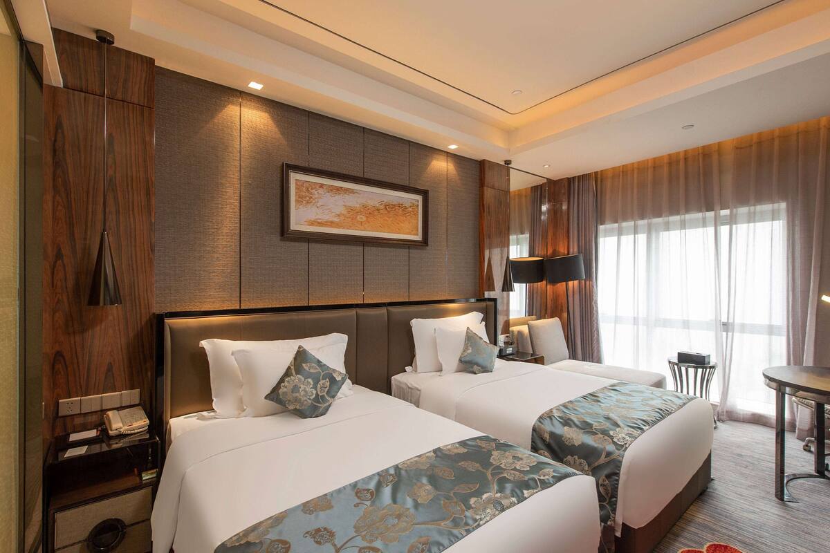 Ein Hotelzimmer mit zwei Betten, einem Nachttisch mit Telefon, einem gerahmten Bild an der Wand und einem Fenster mit Vorhängen.
