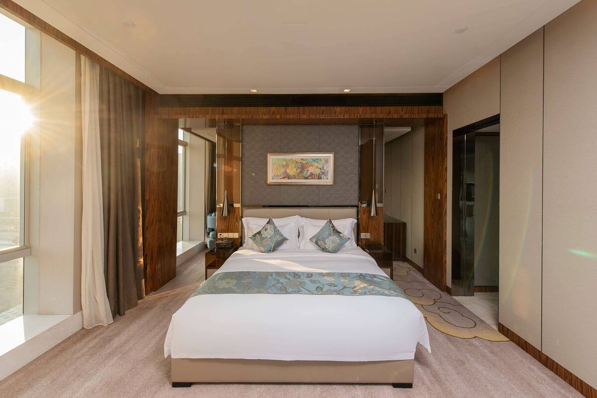 Ein modernes Hotelzimmer mit einem großen Bett, einem Gemälde darüber und Blick auf die Stadt durch das Fenster.