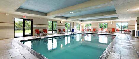 An indoor swimming pool with clear blue water, surrounded by red and blue lounge chairs, and large windows offering views of greenery outside.