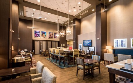 A hotel lobby with a central dining area, multiple seating options, and a large screen displaying a hotel name.