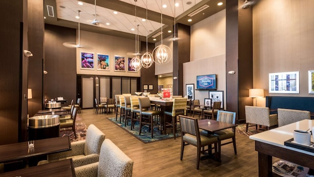 A hotel lobby with a central dining area, multiple seating options, and a large screen displaying a hotel name.