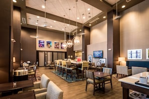 A hotel lobby with a central dining area, multiple seating options, and a large screen displaying a hotel name. - Hampton Inn & Suites Gulfport I-10 (Gulfport)