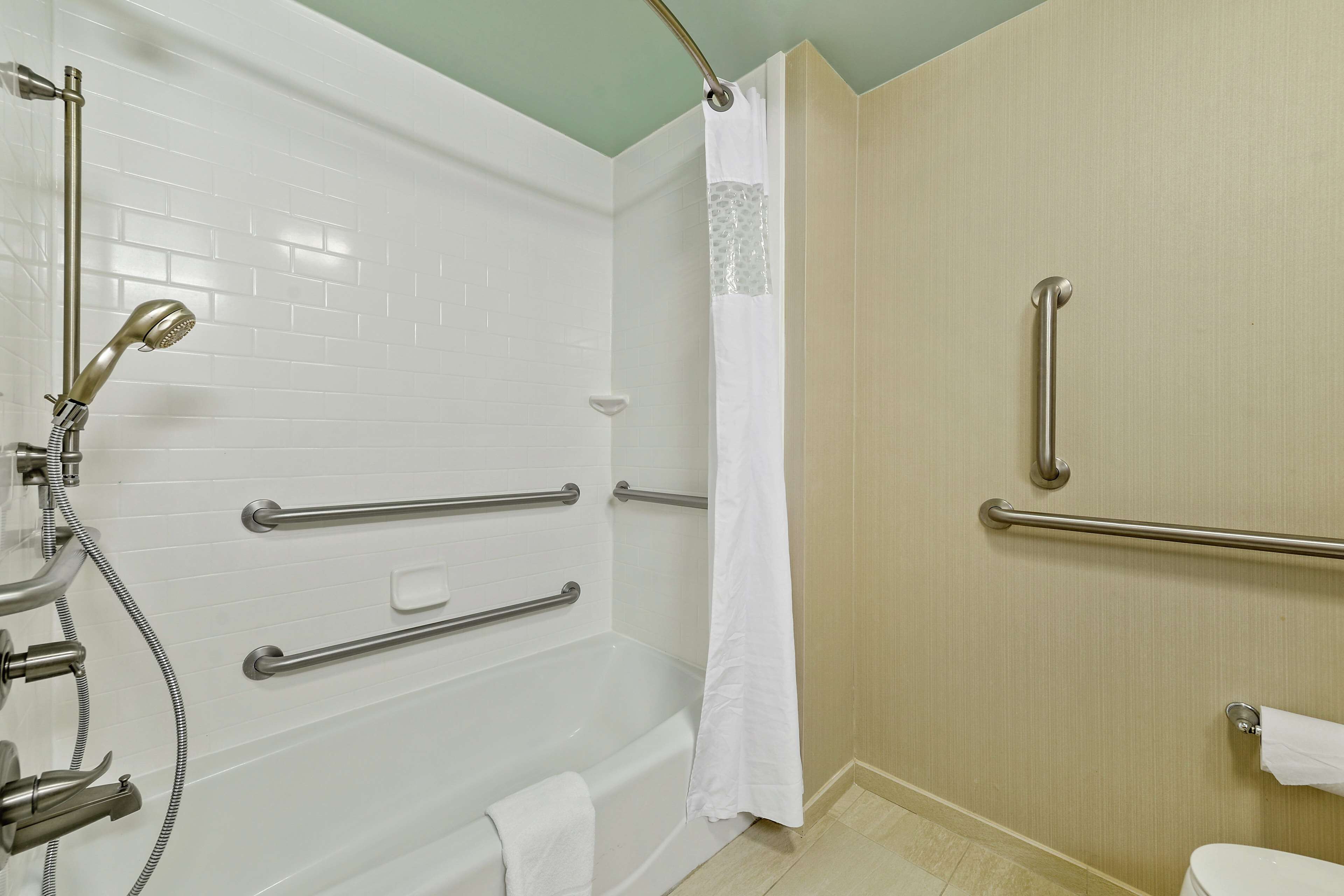 A bathroom with a white bathtub, grab bars, a showerhead, and a toilet.