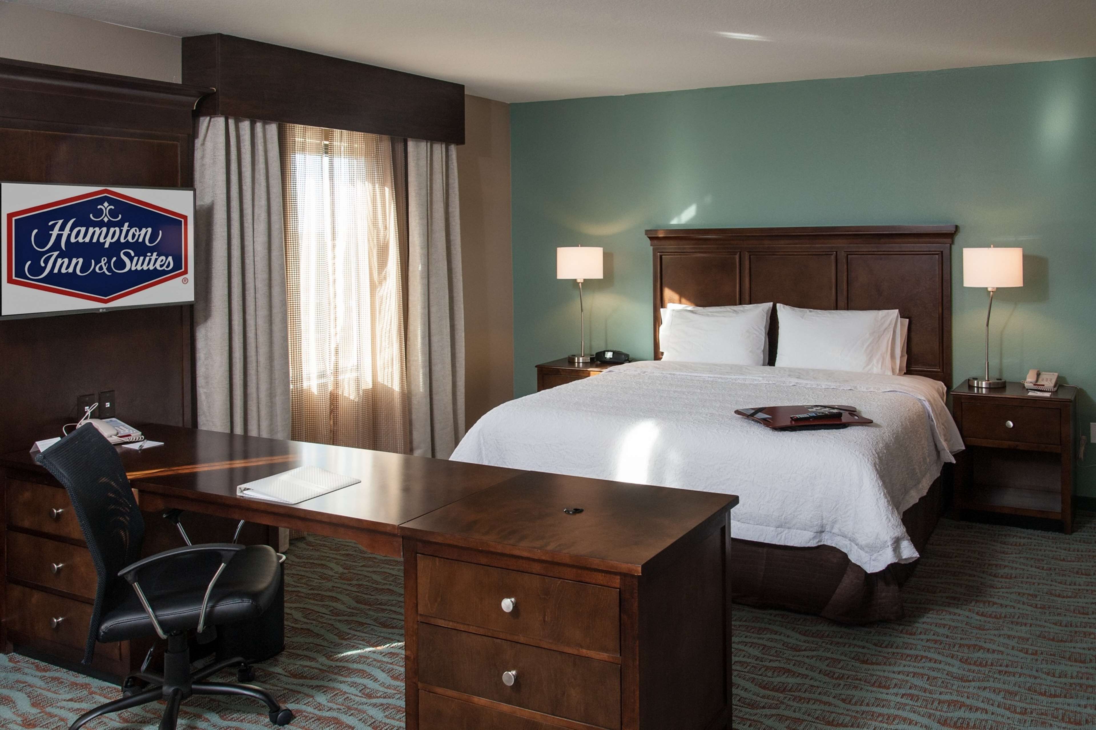 A Hampton Inn & Suites bedroom with a large bed, two bedside lamps, a desk with a chair, and a nightstand with a phone.
