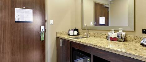 A hotel bathroom with a wooden door, a sink with a mirror, a coffee maker, and a microwave.