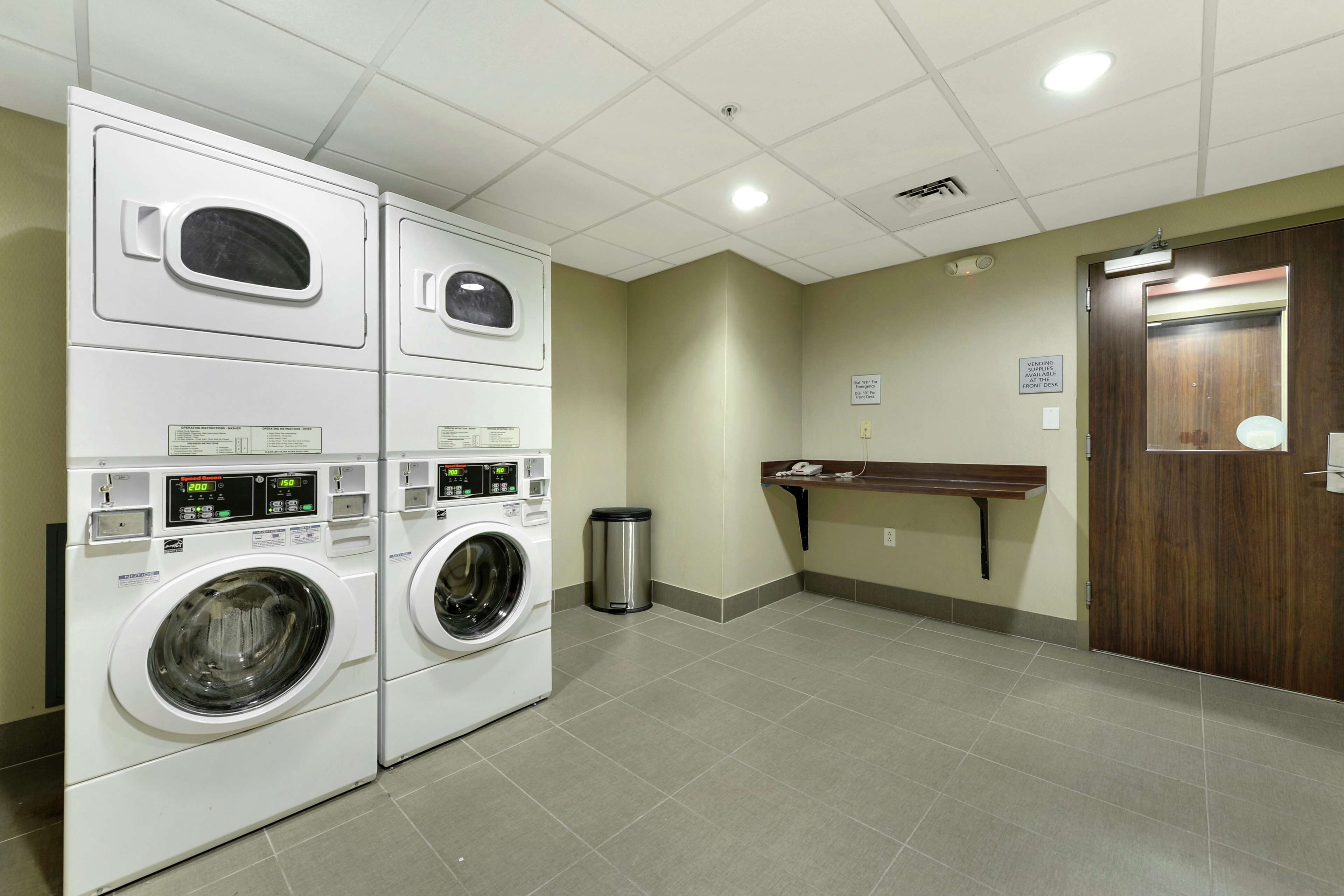 A laundry room with stacked washers and dryers, a trash can, and a wooden bench.