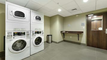A laundry room with stacked washers and dryers, a trash can, and a wooden bench.