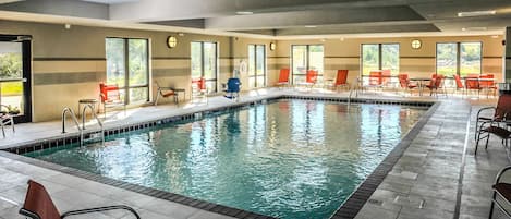 Una piscina interior con agua cristalina, rodeada de sillas y mesas rojas, ventanas amplias y suelo de baldosas.